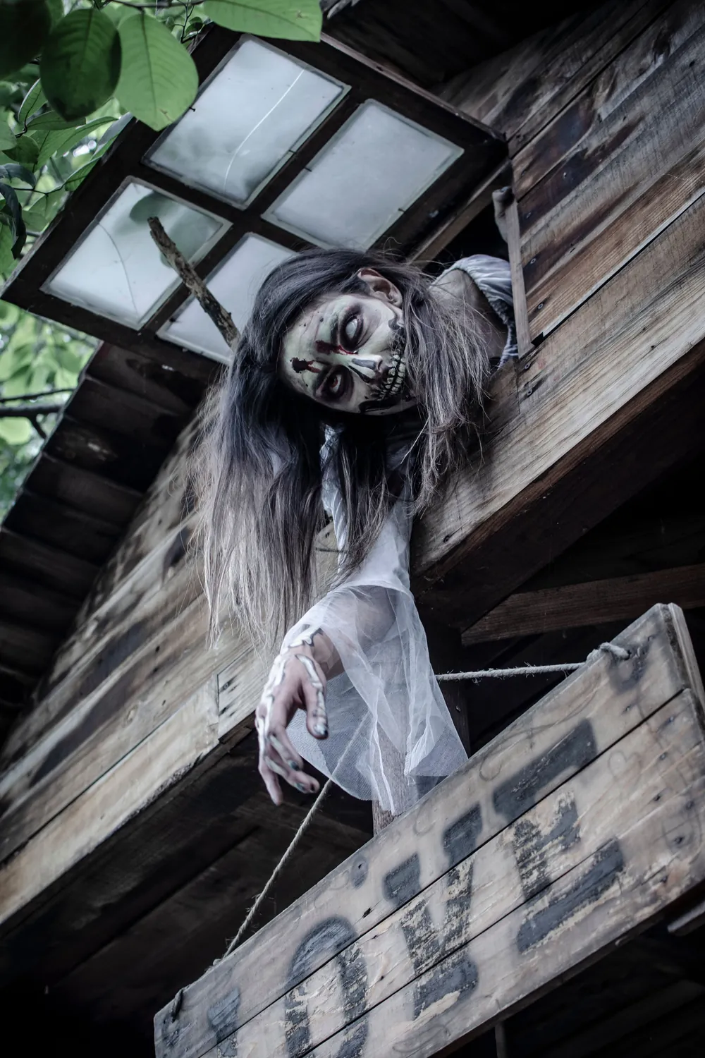A scare actor in zombie makeup leaning out of a wooden treehouse window