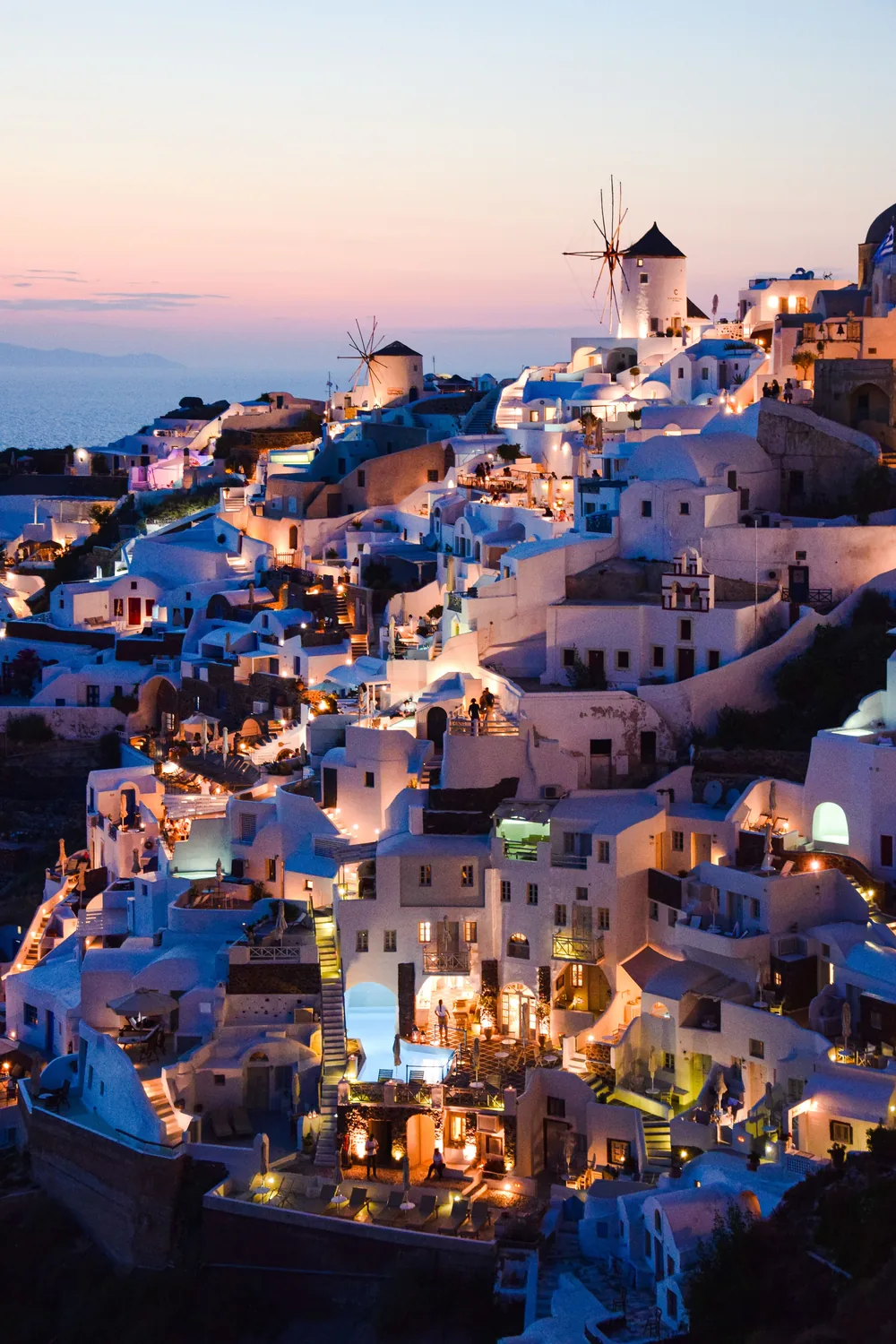 Oia at dusk, its whitewashed houses lit up down the cliff with a windmill silhouetted above