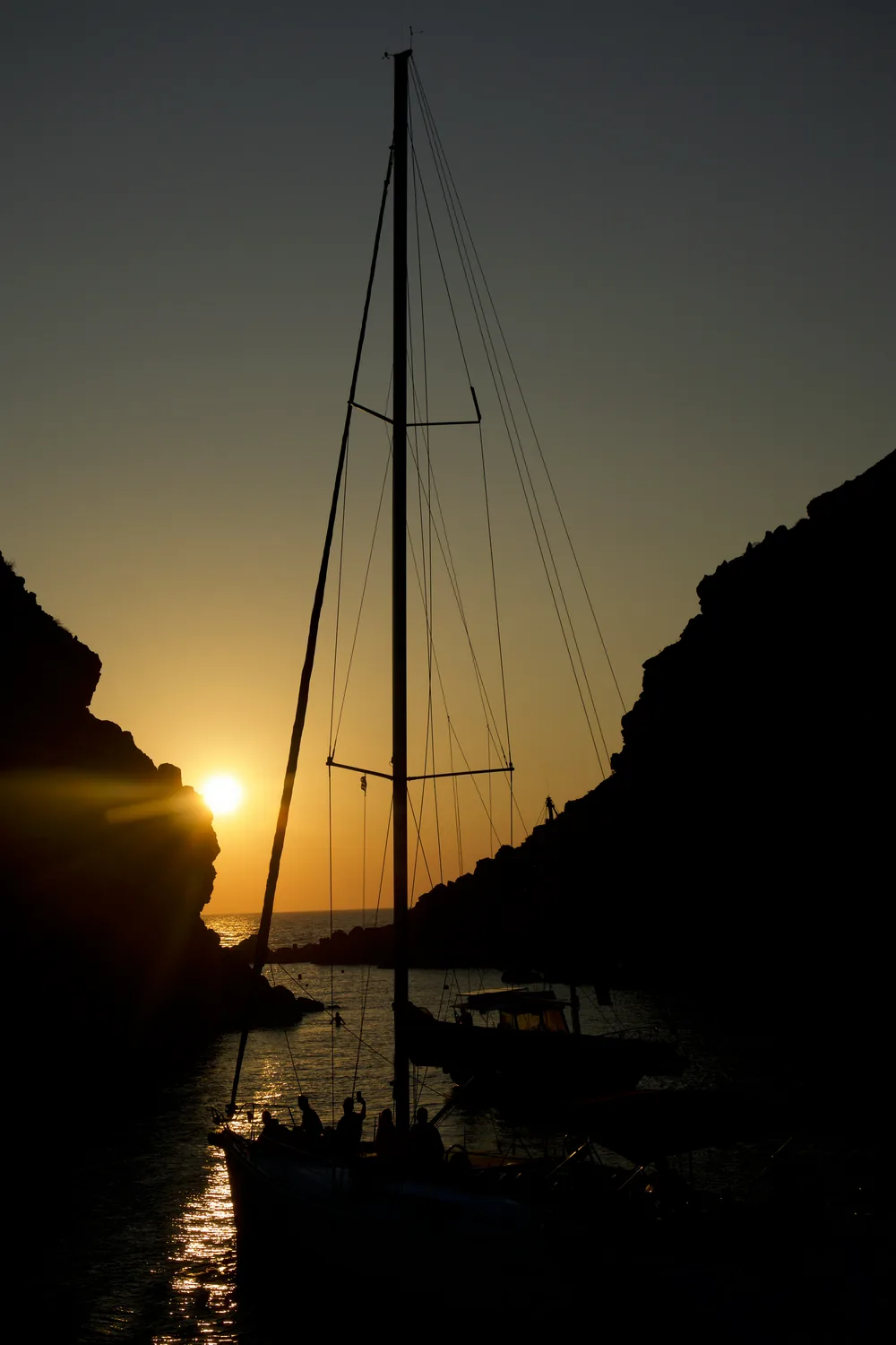 A sailing boat silhouetted in a rocky inlet with the sun setting directly behind its mast