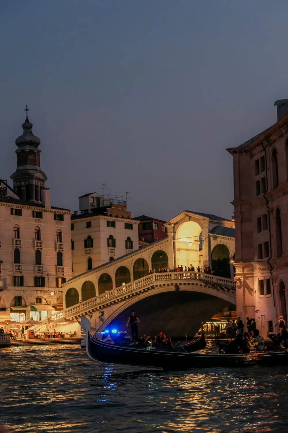The Rialto Bridge at dusk, its arcade lit, with a gondola passing underneath on the Grand Canal