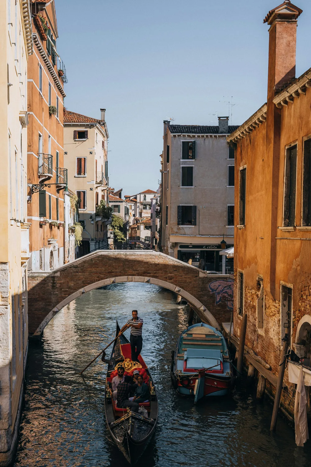 A gondola approaching a low stone bridge on a quiet Venetian side canal, ochre houses on either side