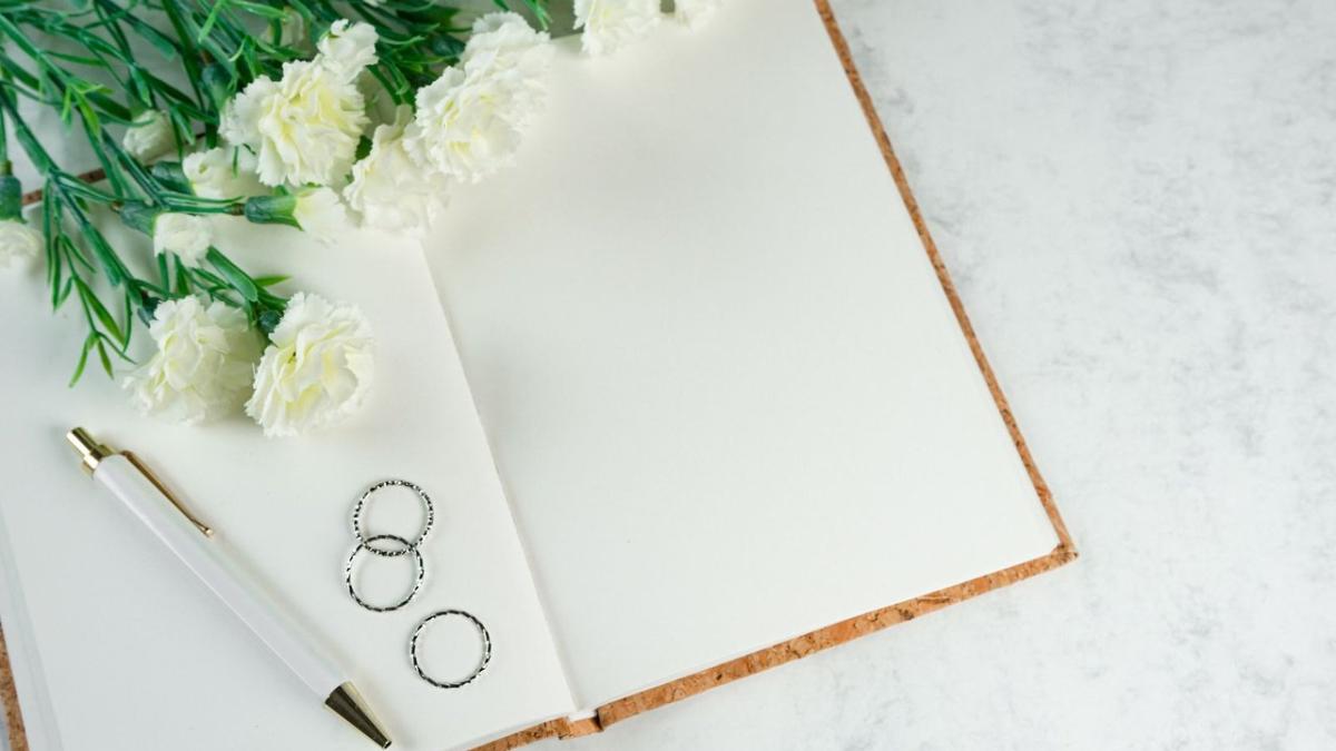 An open notebook, a pen, two rings and white carnations on marble