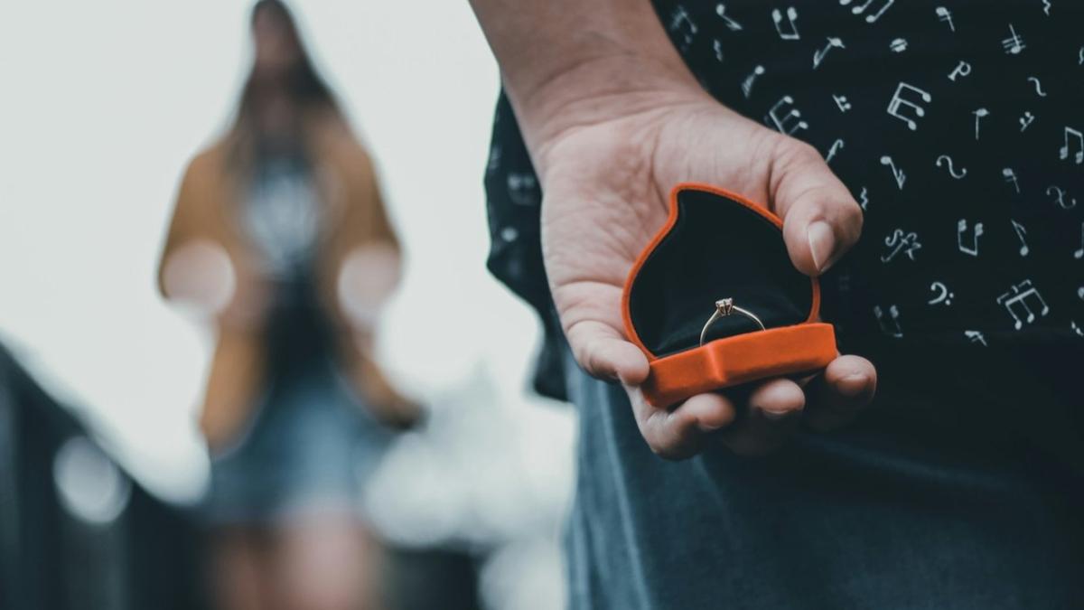 A man holds an open ring box behind his back while she stands out of focus in front of him