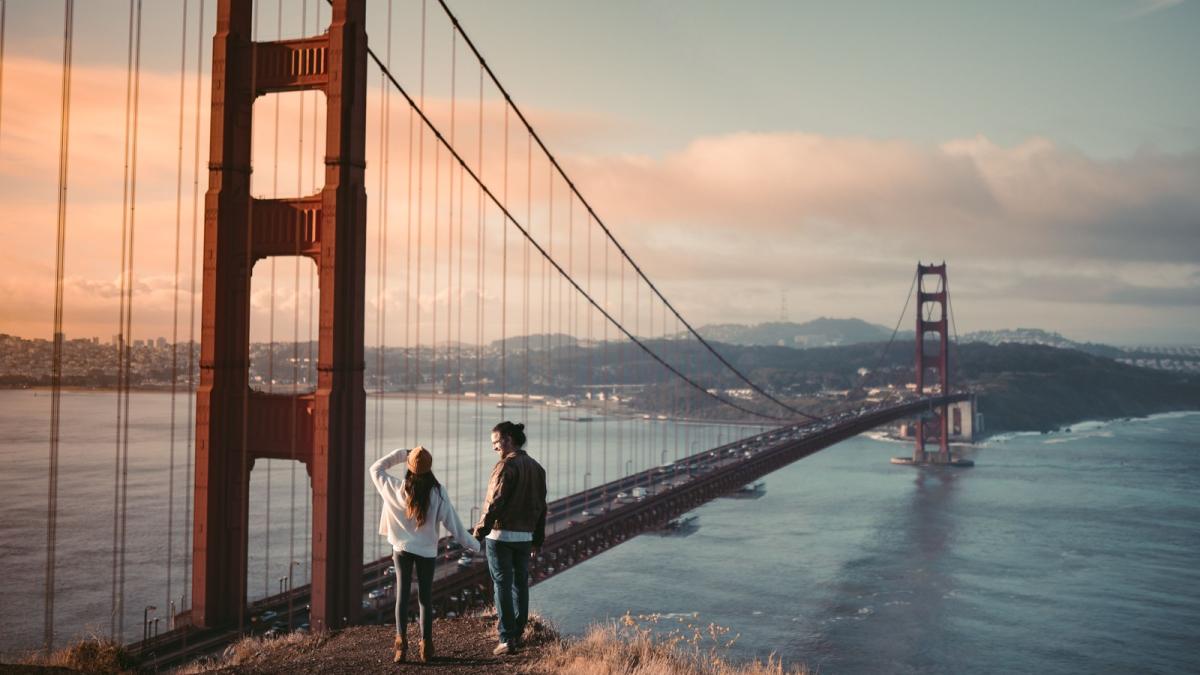 A couple standing on the bluff below the Golden Gate Bridge as the sun rises behind the bay