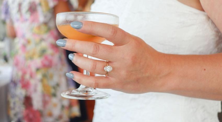A hand wearing an engagement ring holds a cocktail glass at a party