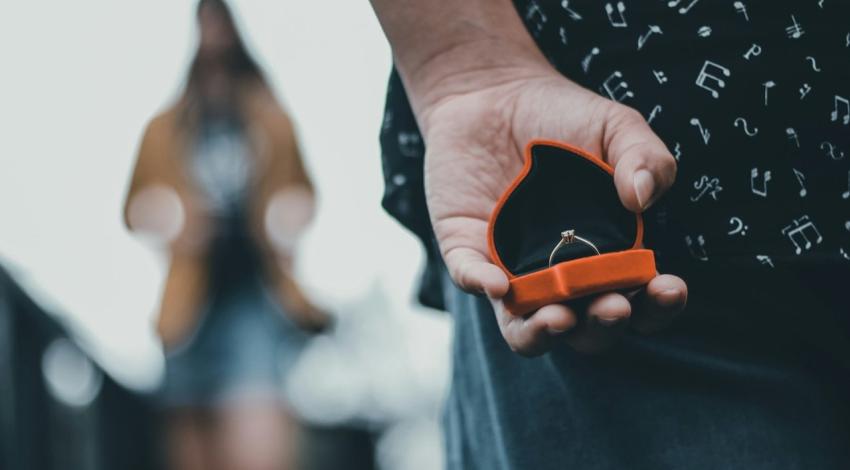 A man holds an open ring box behind his back while she stands out of focus in front of him