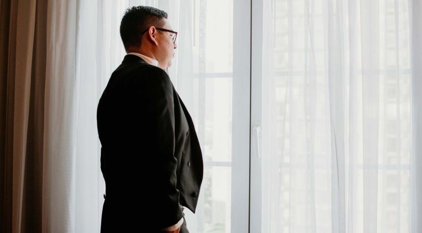 A man in a dark suit looks out of a tall window through sheer curtains