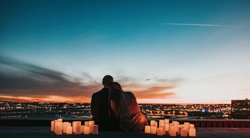 A couple sits among candles on a rooftop, looking out over a city at sunset