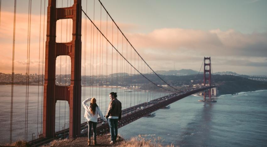 A couple standing on the bluff below the Golden Gate Bridge as the sun rises behind the bay