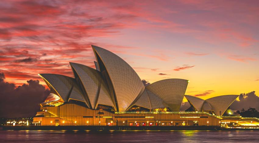 The Sydney Opera House lit up at dusk under a pink and gold sunset sky, seen across the harbour