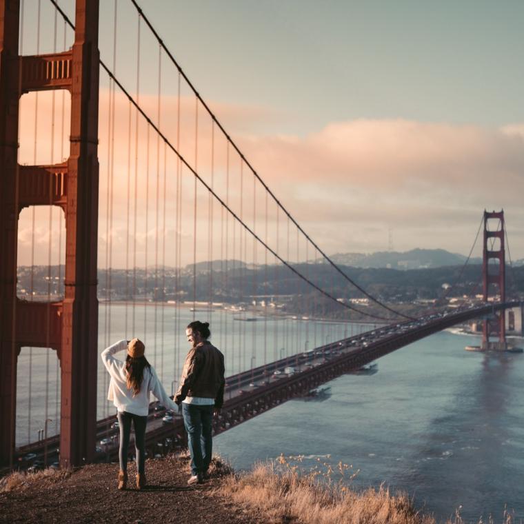 A couple standing on the bluff below the Golden Gate Bridge as the sun rises behind the bay