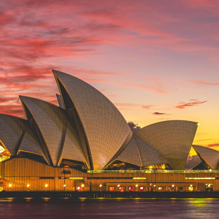 The Sydney Opera House lit up at dusk under a pink and gold sunset sky, seen across the harbour