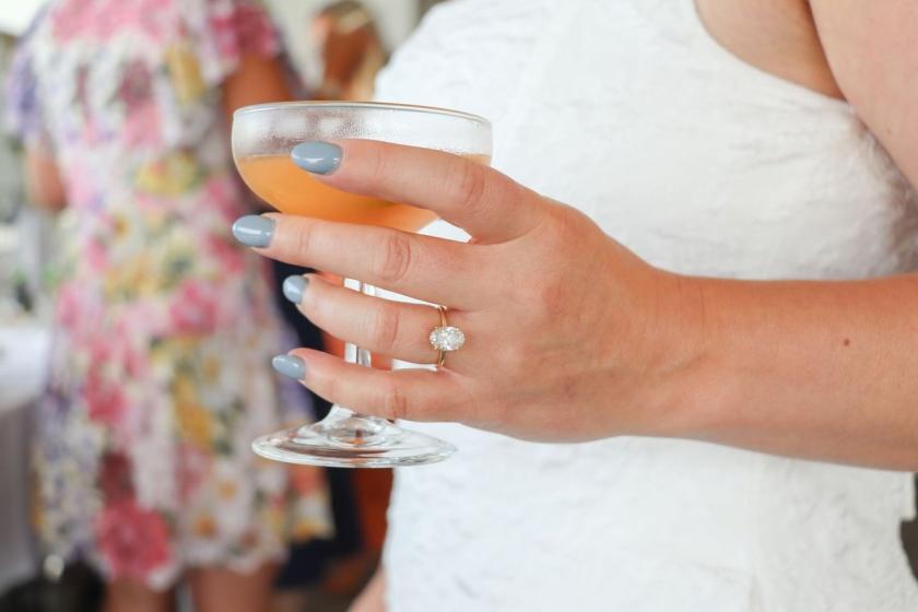 A hand wearing an engagement ring holds a cocktail glass at a party