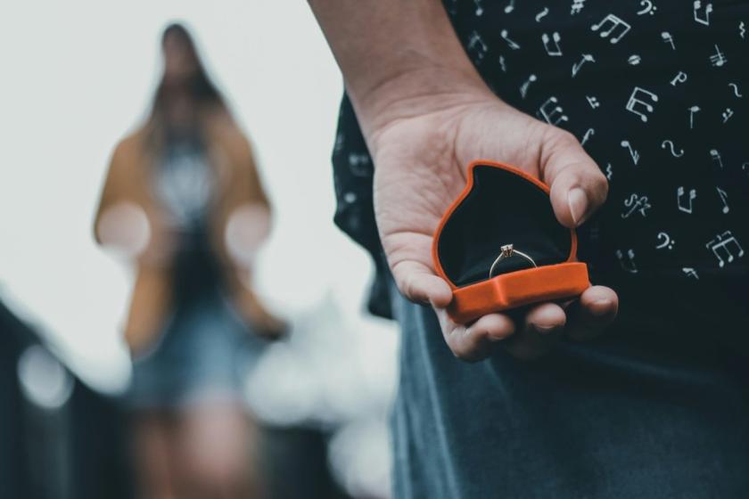 A man holds an open ring box behind his back while she stands out of focus in front of him