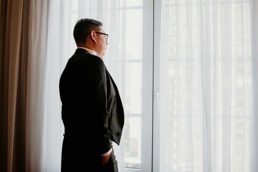 A man in a dark suit looks out of a tall window through sheer curtains