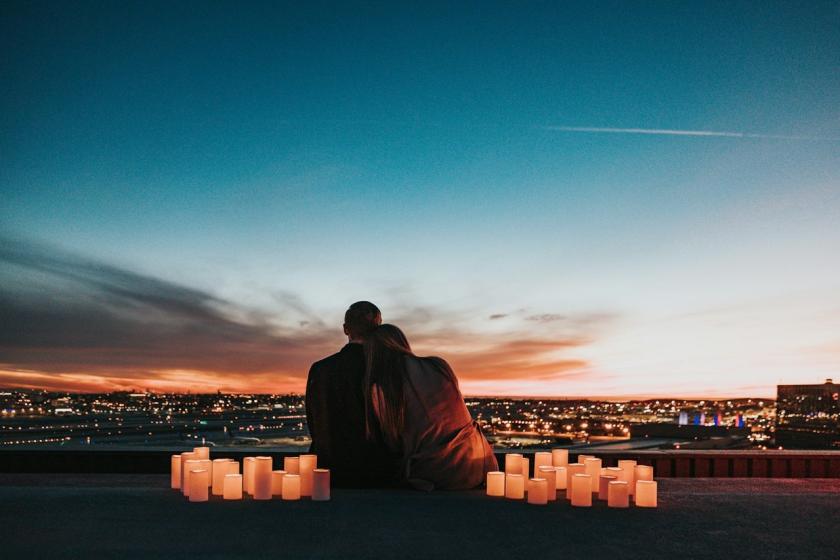 A couple sits among candles on a rooftop, looking out over a city at sunset