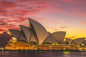 The Sydney Opera House lit up at dusk under a pink and gold sunset sky, seen across the harbour