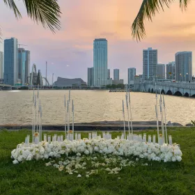 A candlelit white rose installation on a waterfront lawn at sunset, with palm fronds overhead and the Miami skyline across the bay