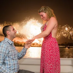A proposal on a boat at night, with fireworks bursting over the water behind the couple
