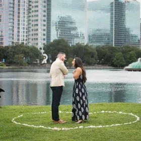 A man and woman face each other inside a circle of rose petals on the lawn at Lake Eola, with the fountain and downtown Orlando towers behind