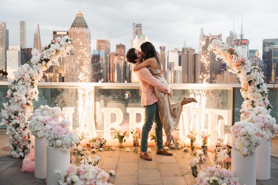 A couple embrace beneath a white and blush floral arch on a rooftop terrace, with illuminated MARRY ME letters and the Manhattan skyline behind them