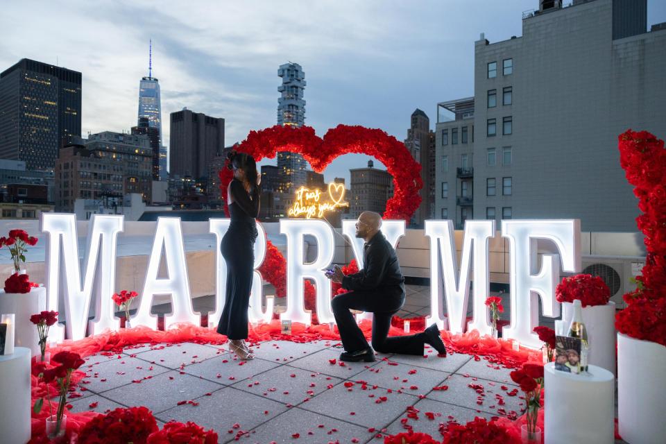 A man kneels to propose inside a heart of red roses, with lit MARRY ME letters and the Lower Manhattan skyline behind