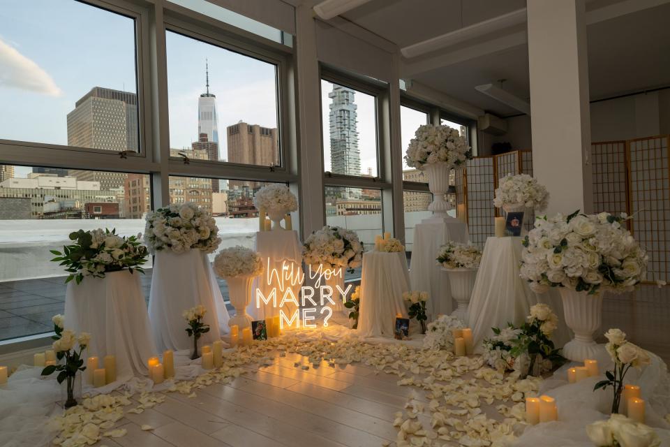 An indoor proposal setup of white roses, candles and a neon Will You Marry Me sign, arranged in front of windows overlooking the city