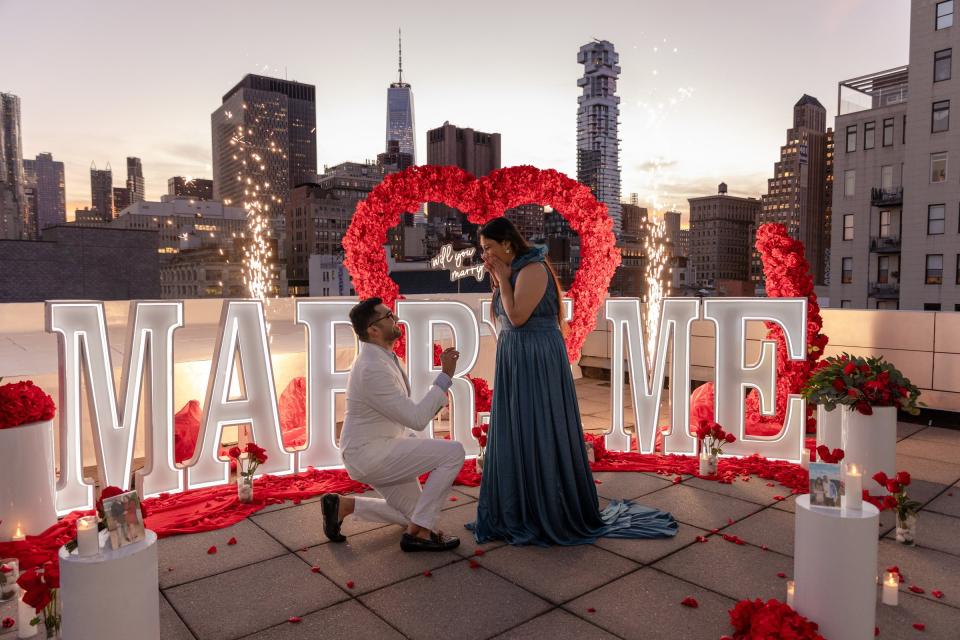 A man in a cream suit kneels to propose as sparklers light up behind a red rose heart and MARRY ME letters on a rooftop at dusk