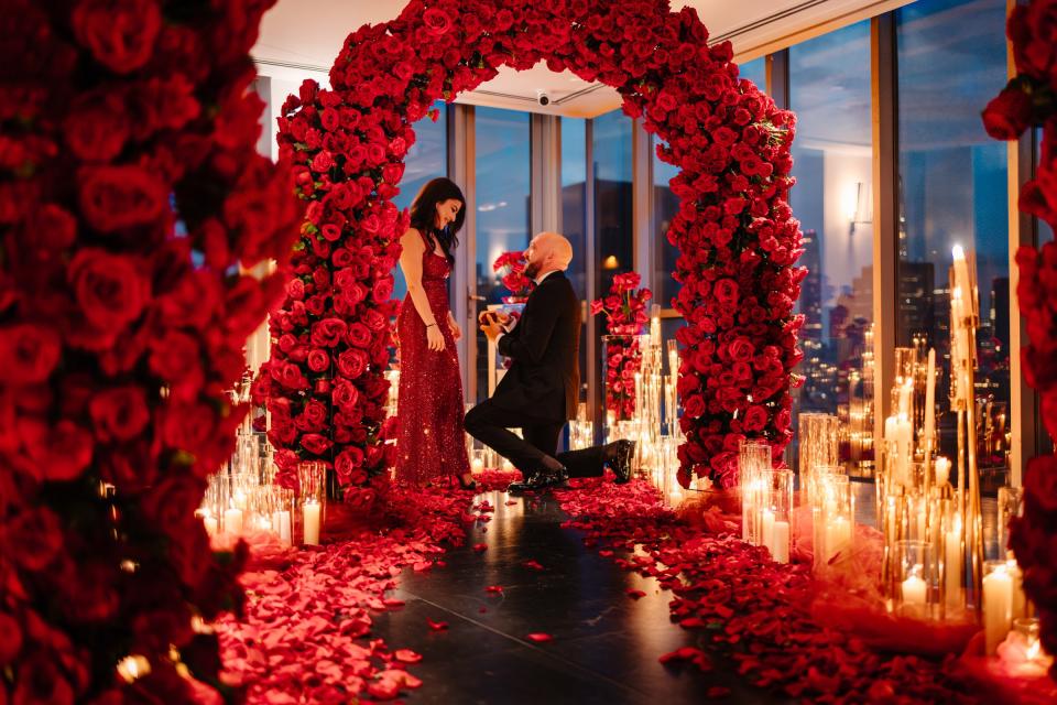 A man kneels to propose beneath a dense arch of red roses, surrounded by tall candles and petals, with the city lit through the windows behind