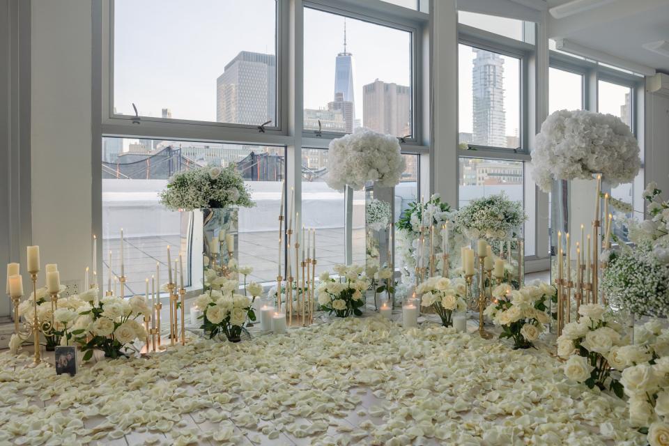 White roses, gold candlesticks and a carpet of petals arranged in front of tall windows overlooking the Brooklyn Bridge