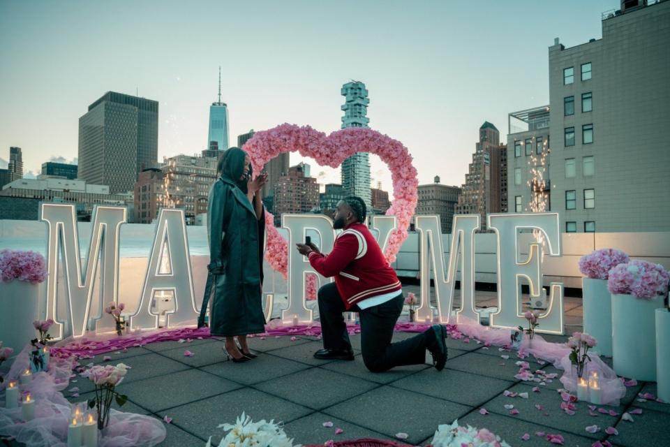 A man kneels to propose in front of a pink rose heart and MARRY ME letters on a rooftop, with the Manhattan skyline behind