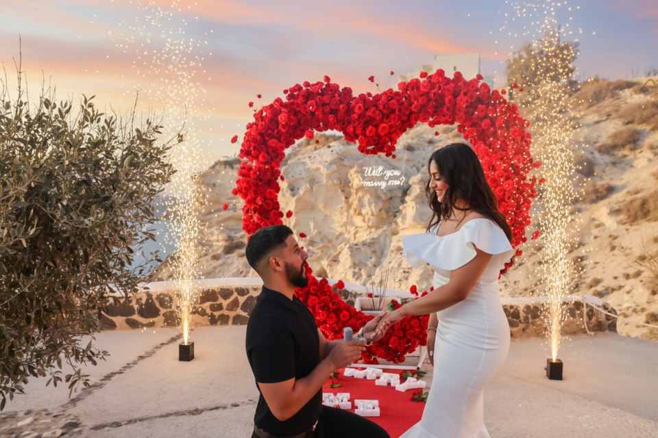 A man kneels to propose to a woman in front of a heart-shaped red rose arch with a neon sign that reads Will you marry me at sunset with fireworks against the breathtaking Santorini caldera. The magical atmosphere, dramatic views, and warm candlelight create an unforgettable proposal moment in one of the world’s most romantic destinations. 