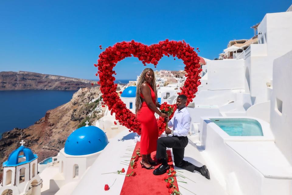 A man in a white shirt and black pants kneels to propose to a woman in a red dress before a large heart-shaped floral arch against the Blue Domes of Santorini.