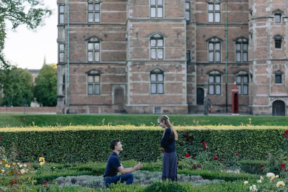A man kneels to propose in the rose garden at Rosenborg Castle in Copenhagen, the castle brickwork and a red sentry box behind them
