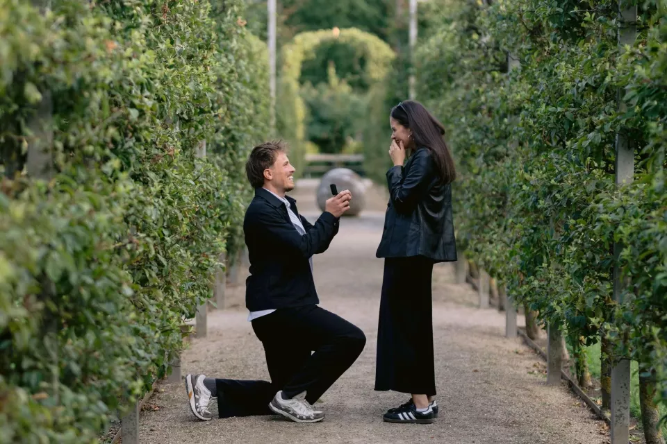 A man proposes on a gravel path between two long rows of espaliered fruit trees, both of them laughing