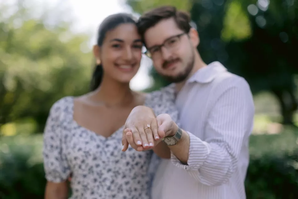 A newly engaged couple hold her hand out to the camera to show the engagement ring, the pair of them soft focused behind it