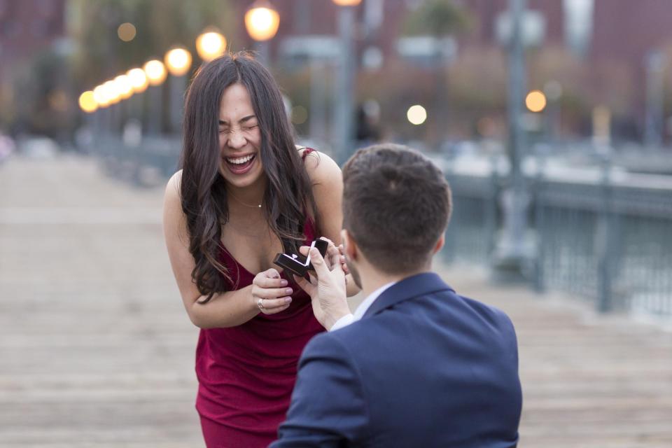 A man kneeling with an open ring box on a lamplit wooden pier at dusk as she laughs with her eyes shut in a burgundy velvet dress