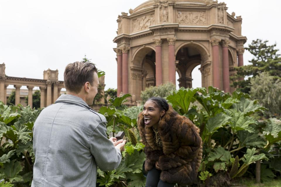 He kneels in a grey blazer with the ring box open as she leans in laughing in a fur coat, the rotunda of the Palace of Fine Arts behind them