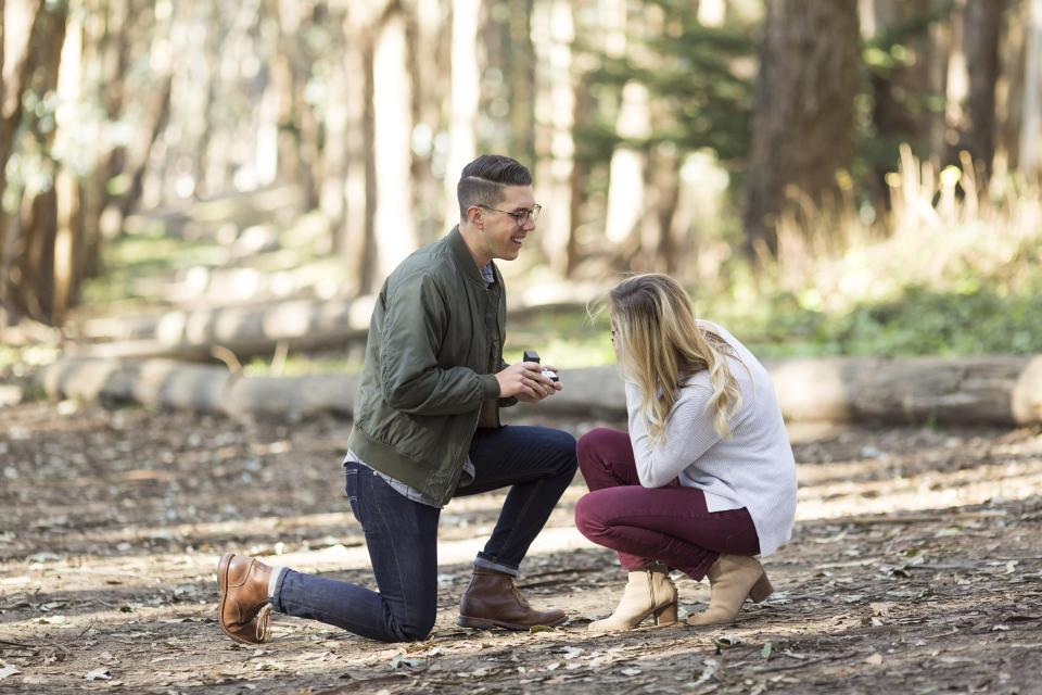 He kneels with the ring box in a sunlit wooded grove as she crouches down opposite him with her hands to her face