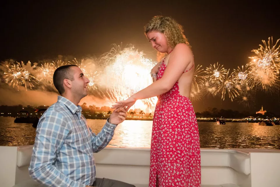 A proposal on a boat at night, with fireworks bursting over the water behind the couple