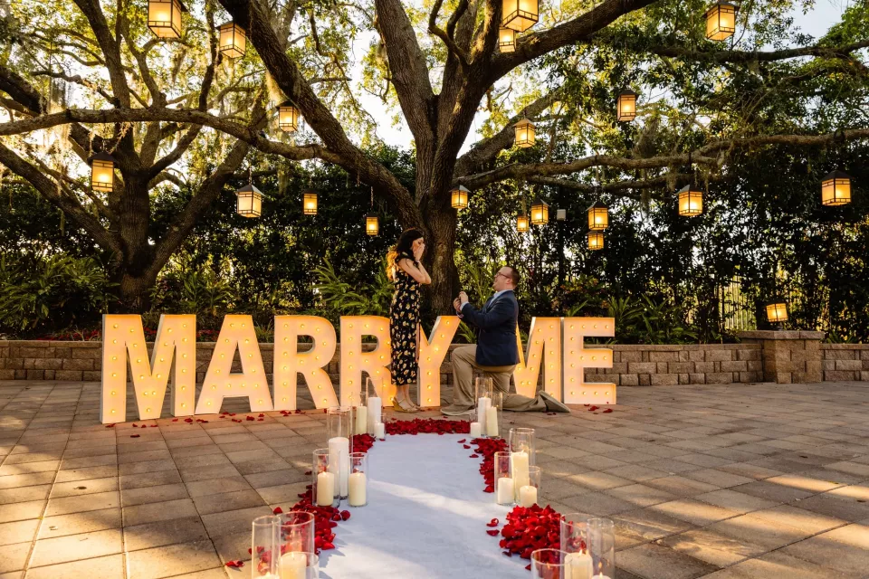 A proposal under a lantern-hung oak tree, behind lit MARRY ME letters and a candlelit aisle of rose petals