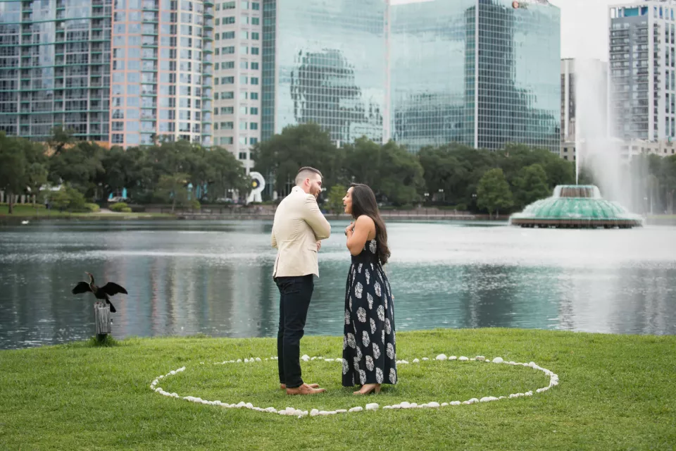 A man and woman face each other inside a circle of rose petals on the lawn at Lake Eola, with the fountain and downtown Orlando towers behind