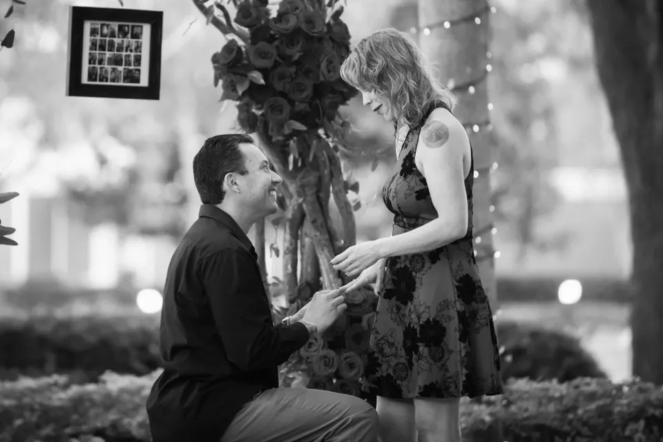 Black and white: he kneels and takes her hand beneath a rose-covered arch strung with fairy lights