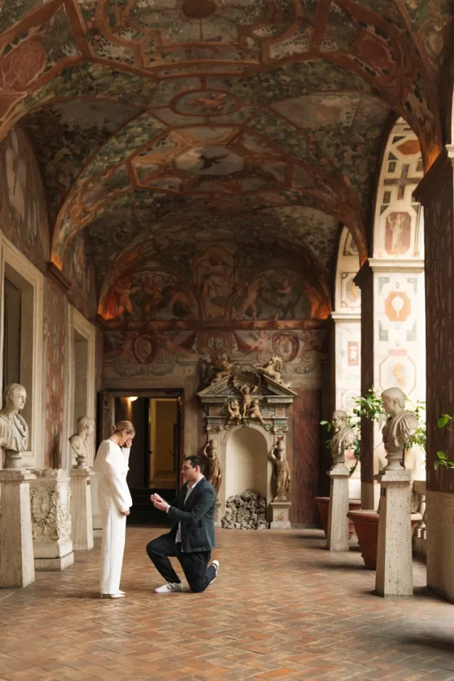 A man on one knee proposing in the frescoed loggia at Palazzo Altemps in Rome, marble busts lining both walls