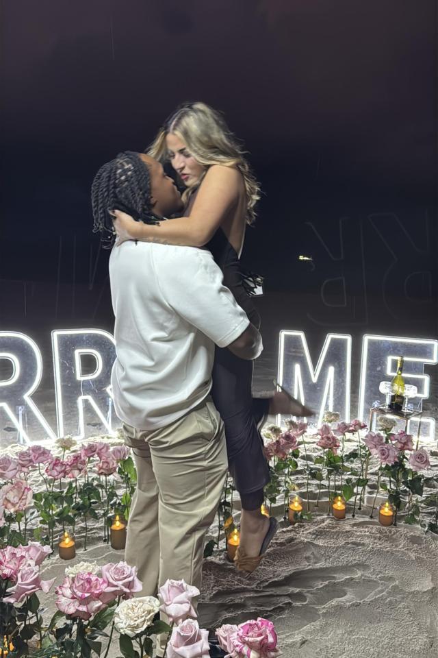 Couple celebrating beside flowers and illuminated MARRY ME letters at night