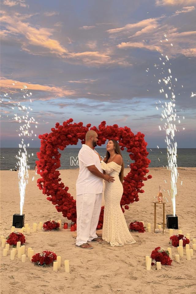 Couple kissing beneath a red floral heart arch at sunset