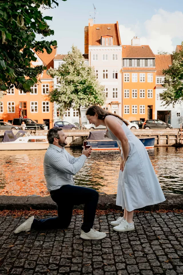 A man on one knee holding out a ring box on a cobbled canal quay in Copenhagen, orange and white townhouses and moored boats across the water