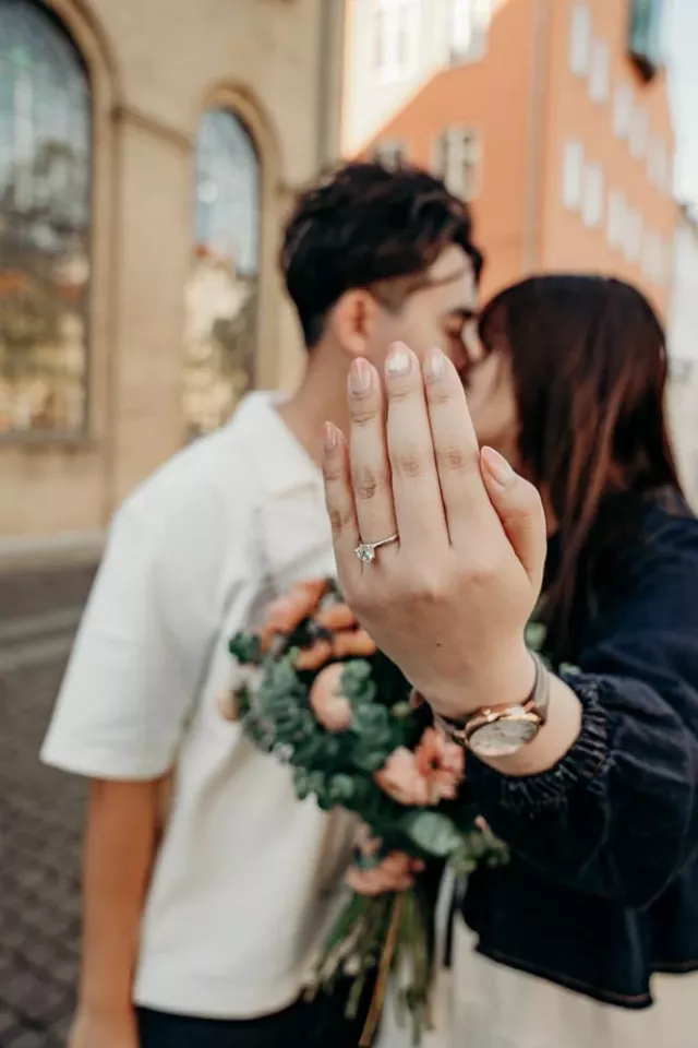 She holds her hand up to the camera to show the solitaire, a bouquet of peach flowers and eucalyptus in her other arm, the couple kissing out of focus behind it