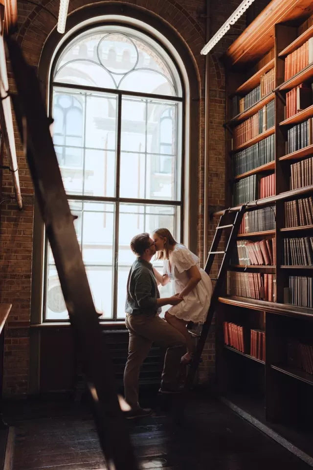 A couple lean in to kiss on a library ladder beside a tall arched window, dark wooden shelves of books filling the wall next to them