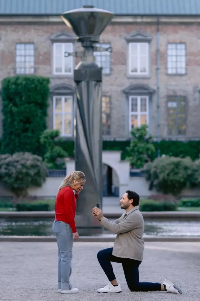 A man on one knee holding out a ring box by the fountain in the Royal Library Garden in Copenhagen, she covers her mouth in surprise